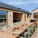 Lange Holztisch mit Stühlen auf Terrasse. Hintergrund: Holzhäuser mit Fenstern und Whirlpool.