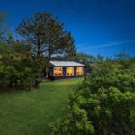 Ein Haus mit großen Fenstern, beleuchtet, umgeben von Gras und Bäumen bei Nacht.