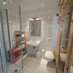 Badezimmer mit Dusche, Waschbecken und Toilette. Holzregal und rosa Wäschekorb.