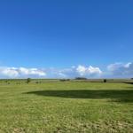 Weite grüne Wiese unter blauem Himmel mit weißen Wolken und schattigen Bereichen.