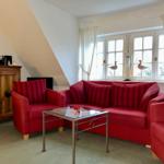 Wohnzimmer mit rotem Sofa, zwei Sesseln und Glas-Tisch. Fenster mit weißen Vorhängen.