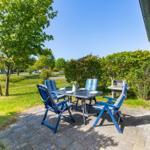 Terrasse mit Tisch, vier Stühlen und Grill. Grünfläche und Bäume im Hintergrund.