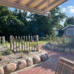 Terrasse mit Sonnenschutz, Sandbeete mit Gräsern und Steinumrandung, Pflasterweg und kleiner Gartenhaus im Hintergrund.