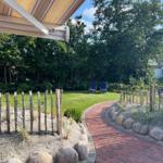 Garten mit Steinterrasse, Holzzaun und Liegestühlen unter Bäumen.