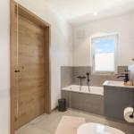 Bad mit Badewanne, Waschbecken und Toilette. Holztür und Fenster mit Jalousie.