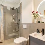 Badezimmer mit Dusche, Waschbecken und Toilette. Rundes Spiegel mit Beleuchtung und Blumen.