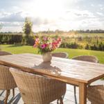 Holztisch mit Stühlen auf Terrasse, Blumenarrangement, Blick auf grünes Feld.