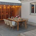 Holztisch mit Stühlen auf Terrasse, beleuchtet durch Girlanden und Laterne.