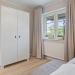Schlafzimmer mit weißem Schrank, Fenster mit Vorhängen und Bett mit weißer Bettwäsche.