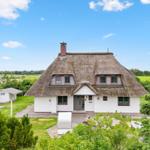Haus mit Strohdach, weiß verputzt, umgeben von Grünflächen und Bäumen unter blauem Himmel.