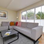 Ein Wohnzimmer mit grauem Sofa, Kissen und Couchtisch. Große Fenster mit Blick auf Garten.