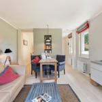 Living room with sofa, dining table, and window. Wooden floor and wall shelf visible.