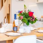 Gedeckter Tisch mit Blumenstrauß und Flasche. Hintergrund: Küchenbereich und Treppe.