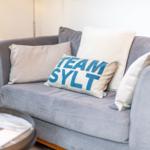 Ein grauer Sofa mit Kissen, eins mit der Aufschrift TEAM SYLT.