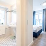Badezimmer mit Fenster und Waschbecken. Nebenraum mit Bett und Vorhängen.