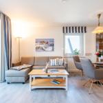 Wohn- und Essbereich mit Sofa, Tisch und Stühlen. Fenster und Bilder an den Wänden.