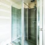 Shower with glass enclosure, dark tiles, and showerhead. Left: heater, right: shelf with mirror.
