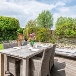 Deck with wooden table, chairs, and lounge chair. Backyard with lawn and hedges.