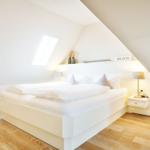 Bedroom with sloped ceiling, white bed, and wooden floor. A nightstand with lamp is next to the bed.