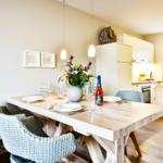 Dining area with wooden table and chairs. Kitchen in background with window.