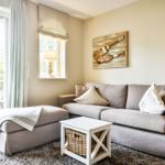 A living room with a gray sofa, ottoman, and white side table. A wooden decoration hangs on the wall.