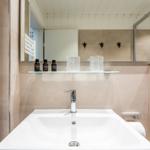 Badezimmer mit Waschbecken, Spiegel und Dusche. Drei Flaschen und zwei Vasen auf der Ablage.