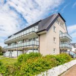 Modern multi-family house with balconies and garden area. White fences and green hedges.