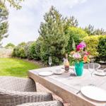 Garten-Terrasse mit gedecktem Tisch, Blumen und Stühlen. Grünflächen und Bäume im Hintergrund.