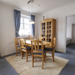 Dining room with wooden table, chairs, and sideboard. Windows with curtains. View into bedroom.