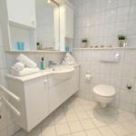 Badezimmer mit Waschbecken, Toilette und Dusche. Weiße Fliesen und Handtücher.