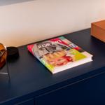 Ein Buch über Pop Art liegt auf einem dunkelblauen Schrank neben einer braunen Schachtel und einer Glaslaterne.