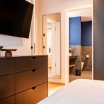 Schlafzimmer mit Bett, Kleiderschrank und Fernseher. Badezimmer mit blauer Wand sichtbar.