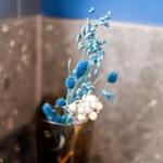 Blauer Blumenschmuck in einem Glas auf einer Wand mit grauen Fliesen.