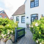Haus mit weißer Fassade, dunklen Fenstern und gepflastertem Weg durch grüne Hecken.