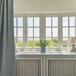 Grosses Fenster mit Blick auf Grünland und Meer. Auf Fensterbank stehen zwei Lampen und eine Pflanze.