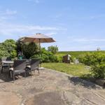 Terrasse mit Tisch, Stühlen und Sonnenschirm. Steinplatten, Gras und Sträucher im Hintergrund.