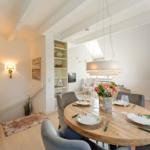 Dining area with round table, gray chairs, and floral decoration. Kitchen and living area are visible.