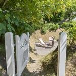 A white gate leads to a terrace with a lounge chair and table under trees.