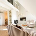 Living room with sofa, armchair, and TV under sloped ceilings. Kitchen and dining area are visible.