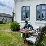 Haus mit weißer Fassade und blauen Fenstern. Auf dem Grundstück steht eine Figur am Tisch.