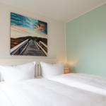 Schlafzimmer mit weißem Bett, Wandbild und Nachttisch mit Lampe.