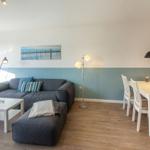 Wohn- und Essbereich mit grauem Sofa, weißem Tisch und Stühlen, blau-weißen Wänden und Holzboden.