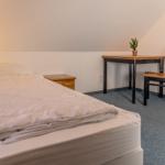 Ein Schlafzimmer mit Bett, Nachttisch, Tisch und Stuhl. An der Wand hängt eine Lampe.