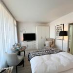 Schlafzimmer mit Bett, TV, Schrank und Sitzbereich. Fenster mit weißen Vorhängen.