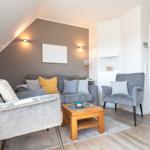 Wohnzimmer mit grauer Couch, zwei Sesseln und Holztisch. Wandfarbe grau, Holzboden und weiße Türen.