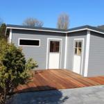 Moderne graue Gartenhütte mit weißer Tür und Holzterrasse. Hintergrund mit Bäumen und klarem Himmel.