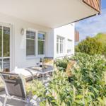 Terrasse mit Tisch, Stühlen und Weinflasche. Umgeben von grünen Pflanzen und blauem Himmel.