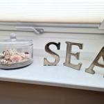 Auf der Fensterbank steht ein Glasbehälter mit Muscheln und Sand neben den Buchstaben SEA.