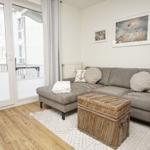 Ein Wohnzimmer mit grauem Sofa, Kissen und einem Korbtablett. Durchgehende Glastür zum Balkon.