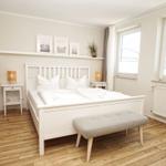 Schlafzimmer mit weißem Bett, Holzboden und Fenstern. An der Wand hängen Bilder.